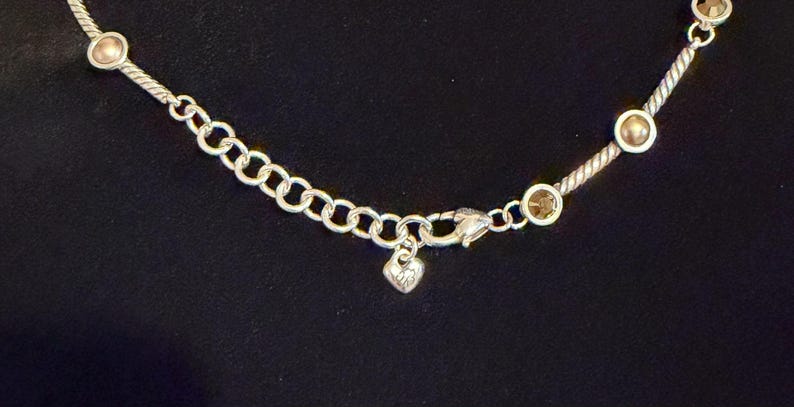 May include: Silver-toned necklace with a delicate chain and a heart-shaped charm. The necklace features small, round, gold-colored accents and a lobster clasp closure. The background is a solid black.