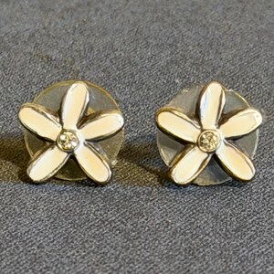 May include: A pair of flower-shaped stud earrings. Each earring has a white enamel flower with five petals and a small, sparkling centre stone. The earrings are set on a silver-coloured base, creating a delicate and feminine design.