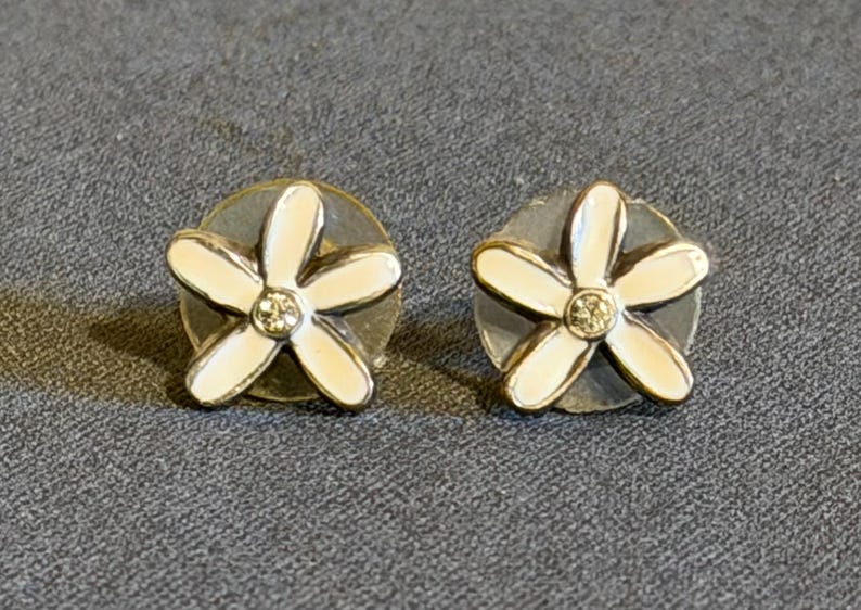May include: A pair of flower-shaped stud earrings. Each earring has a white enamel flower with five petals and a small, sparkling centre stone. The earrings are set on a silver-coloured base, creating a delicate and feminine design.