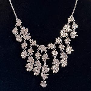 May include: Silver necklace with a cascade of small, daisy-shaped flower pendants. The necklace has a delicate chain and a bib-style design, with the flowers arranged in a layered pattern. The background is a solid black.