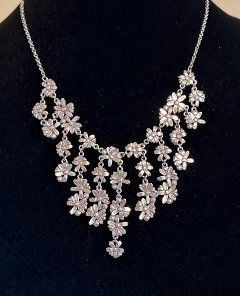 May include: Silver necklace with a cascade of small, daisy-shaped flower pendants. The necklace has a delicate chain and a bib-style design, with the flowers arranged in a layered pattern. The background is a solid black.