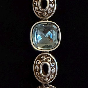 May include: A silver bracelet featuring alternating light blue, square-cut gemstones and decorative, circular silver links. The gemstones are faceted, catching the light. The background is a plain black.