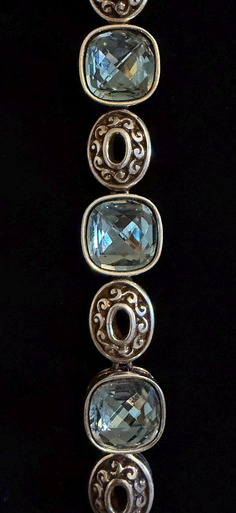 May include: A silver bracelet featuring alternating light blue, square-cut gemstones and decorative, circular silver links. The gemstones are faceted, catching the light. The background is a plain black.