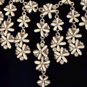 May include: A silver necklace with white enamel flower pendants and crystal details. The floral design is linked by a fine chain, creating a charming and elegant accessory. The necklace is set against a dark background.