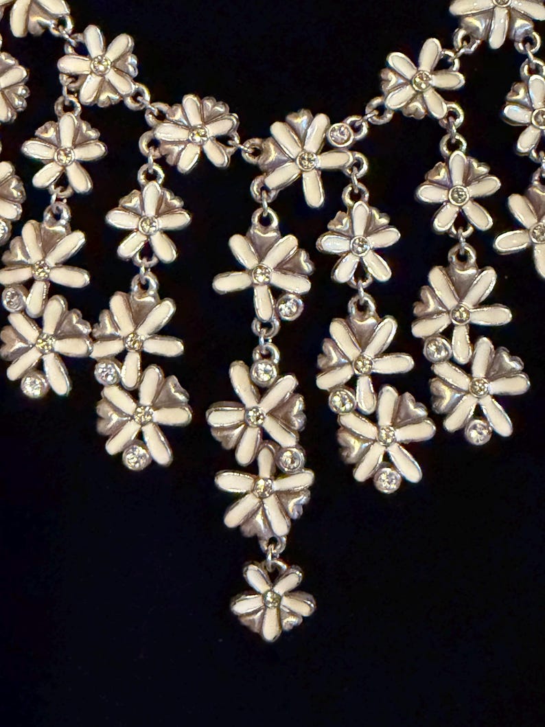 May include: A silver necklace with white enamel flower pendants and crystal details. The floral design is linked by a fine chain, creating a charming and elegant accessory. The necklace is set against a dark background.