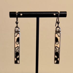 K&ouml;nnte beinhalten: Ein Paar silberfarbene H&auml;ngeohrringe. Jeder Ohrring hat ein rechteckiges Design mit cremefarbenem Hintergrund und schwarzen und grauen abstrakten Formen. Die Ohrringe werden auf einem schwarzen Ohrringst&auml;nder pr&auml;sentiert.