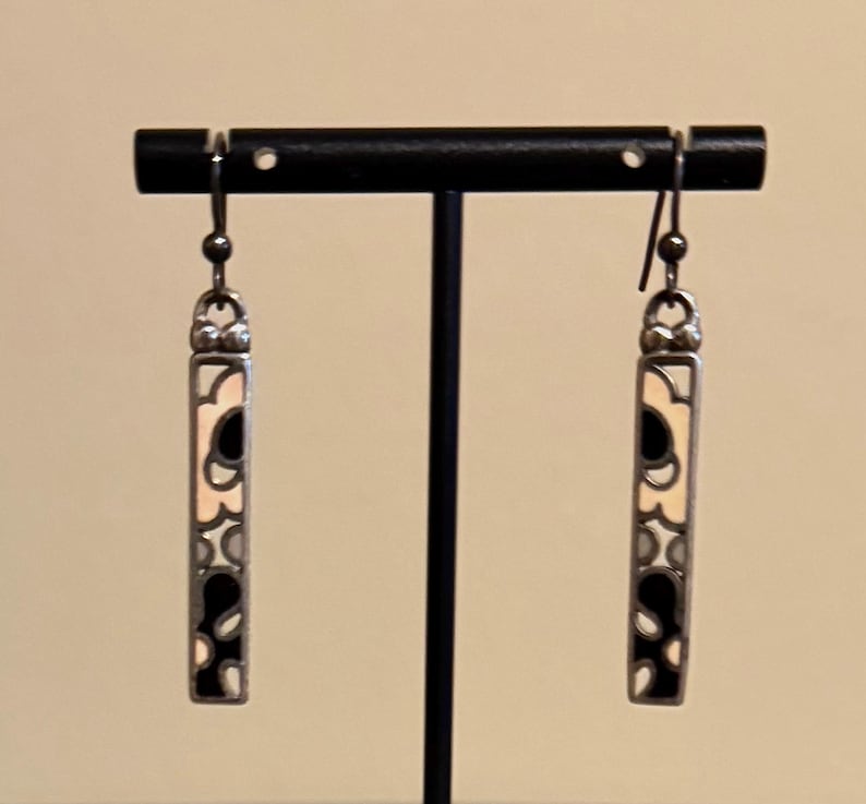 K&ouml;nnte beinhalten: Ein Paar silberfarbene H&auml;ngeohrringe. Jeder Ohrring hat ein rechteckiges Design mit cremefarbenem Hintergrund und schwarzen und grauen abstrakten Formen. Die Ohrringe werden auf einem schwarzen Ohrringst&auml;nder pr&auml;sentiert.