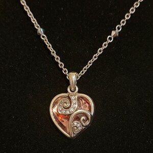 May include: Silver-toned necklace with a heart-shaped pendant. The pendant features a pink-orange center with a swirling design and small clear crystals. The chain is delicate and has small, faceted beads.