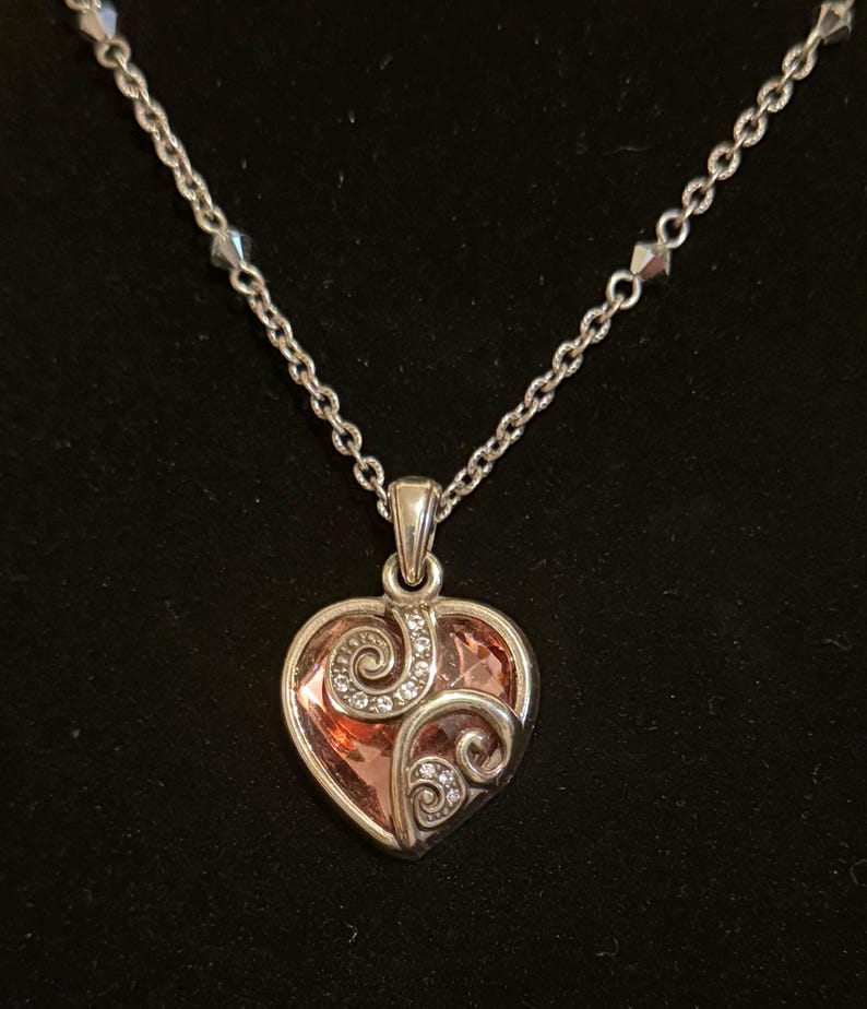 May include: Silver-toned necklace with a heart-shaped pendant. The pendant features a pink-orange center with a swirling design and small clear crystals. The chain is delicate and has small, faceted beads.