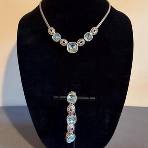 May include: A silver-coloured necklace and bracelet set. The necklace features a series of light blue square and oval-shaped stones, with a matching bracelet. The jewellery is displayed on a black mannequin.