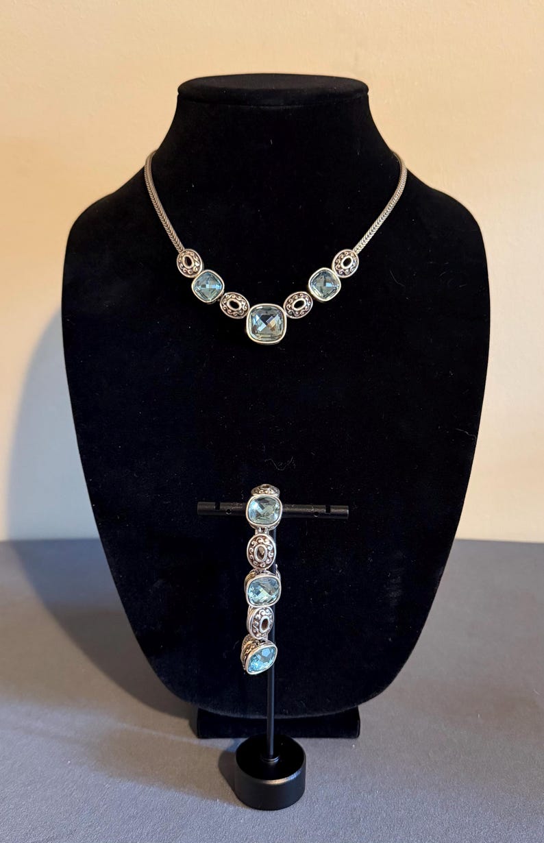 May include: A silver-coloured necklace and bracelet set. The necklace features a series of light blue square and oval-shaped stones, with a matching bracelet. The jewellery is displayed on a black mannequin.