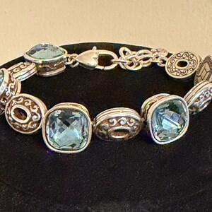 May include: Silver bracelet with square, pale blue gemstones and decorative round and oval links. The bracelet has a lobster clasp. The gemstones are set in silver-toned metal with ornate detailing.