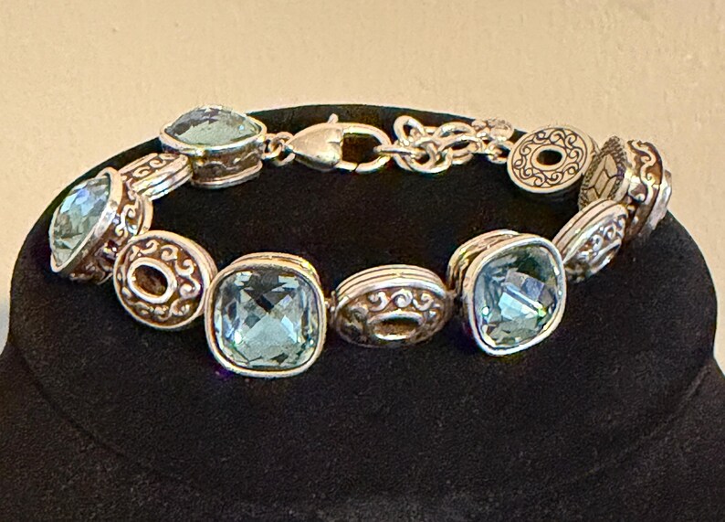 May include: Silver bracelet with square, pale blue gemstones and decorative round and oval links. The bracelet has a lobster clasp. The gemstones are set in silver-toned metal with ornate detailing.