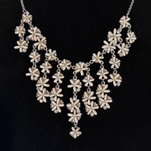 May include: A silver necklace featuring a series of white and silver flower pendants. Each flower has a small, yellow centre and is embellished with clear crystals. The necklace is set against a black backdrop.