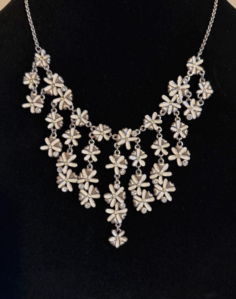 May include: A silver necklace featuring a series of white and silver flower pendants. Each flower has a small, yellow centre and is embellished with clear crystals. The necklace is set against a black backdrop.