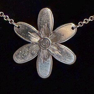 Może przedstawiać: Srebrny naszyjnik z wisiorkiem w kształcie stokrotki. Wisiorek ma sześć płatk&oacute;w i szczeg&oacute;łowy środek ze spiralnym wzorem. Łańcuszek jest delikatny, a tło czarne.