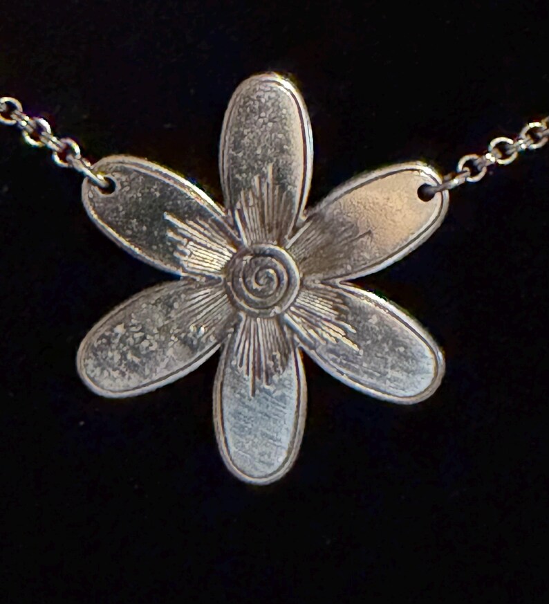 Może przedstawiać: Srebrny naszyjnik z wisiorkiem w kształcie stokrotki. Wisiorek ma sześć płatk&oacute;w i szczeg&oacute;łowy środek ze spiralnym wzorem. Łańcuszek jest delikatny, a tło czarne.