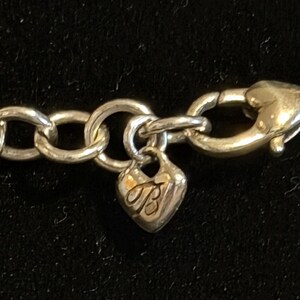 May include: Close-up of a silver-toned necklace clasp and chain. The clasp is a lobster claw style. A heart-shaped charm with the letters "TB" is attached to the chain. The chain links are large and circular.