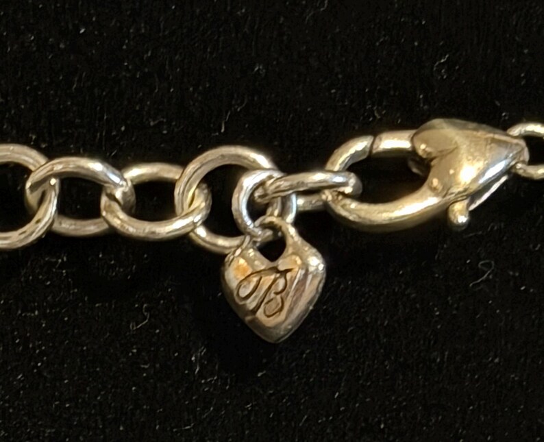 May include: Close-up of a silver-toned necklace clasp and chain. The clasp is a lobster claw style. A heart-shaped charm with the letters "TB" is attached to the chain. The chain links are large and circular.