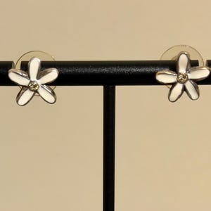 May include: A pair of flower-shaped stud earrings. Each earring has a white enamel flower design with five petals and a small, clear gemstone in the centre. They are displayed on a black jewellery stand.
