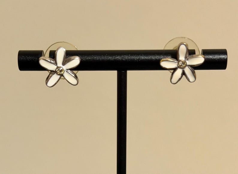 May include: A pair of flower-shaped stud earrings. Each earring has a white enamel flower design with five petals and a small, clear gemstone in the centre. They are displayed on a black jewellery stand.