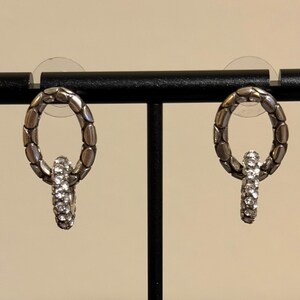 Puede incluir: Un par de pendientes plateados. Cada pendiente presenta un aro ovalado con una superficie texturizada y un elemento m&aacute;s peque&ntilde;o, colgante, con incrustaciones de cristal. Los pendientes se exhiben en un soporte negro sobre un fondo neutro.