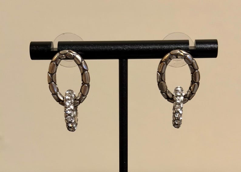 Puede incluir: Un par de pendientes plateados. Cada pendiente presenta un aro ovalado con una superficie texturizada y un elemento m&aacute;s peque&ntilde;o, colgante, con incrustaciones de cristal. Los pendientes se exhiben en un soporte negro sobre un fondo neutro.