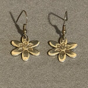 Może przedstawiać: Para srebrnych kolczyk&oacute;w w kształcie kwiat&oacute;w w odcieniu srebra. Każdy kolczyk ma sześciopłatkowy wz&oacute;r kwiatu ze szczeg&oacute;łowym środkiem i haczykiem do przekłutych uszu. Kolczyki są prezentowane na szarej powierzchni.