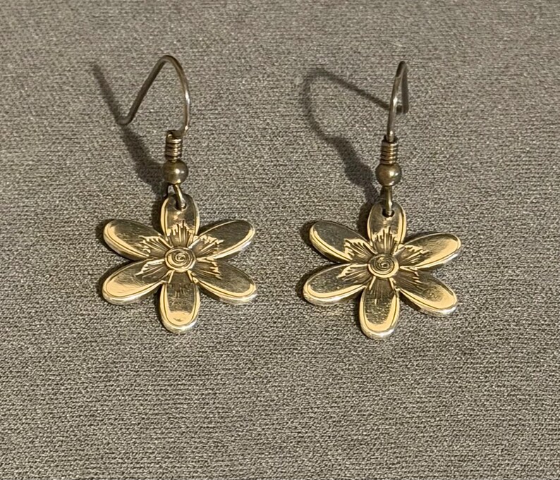 Może przedstawiać: Para srebrnych kolczyk&oacute;w w kształcie kwiat&oacute;w w odcieniu srebra. Każdy kolczyk ma sześciopłatkowy wz&oacute;r kwiatu ze szczeg&oacute;łowym środkiem i haczykiem do przekłutych uszu. Kolczyki są prezentowane na szarej powierzchni.