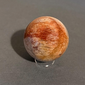 May include: A polished, spherical stone with a mix of reddish-brown and tan hues. The orb is displayed on a clear, circular stand, set against a dark gray background. The stone's surface has a wood-like grain pattern.