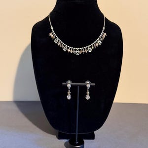 May include: A silver-toned necklace and matching earrings set. The necklace features small, decorative elements and dangling beads. The earrings have a similar design, with a dangling bead. The jewelry is displayed on a black mannequin.