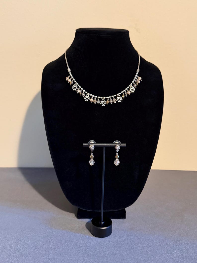 May include: A silver-toned necklace and matching earrings set. The necklace features small, decorative elements and dangling beads. The earrings have a similar design, with a dangling bead. The jewelry is displayed on a black mannequin.