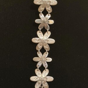 Może przedstawiać: Srebrna bransoletka z ogniwami w kształcie stokrotek. Każdy kwiat ma szczeg&oacute;łowe wzory płatk&oacute;w i środka. Bransoletka jest prezentowana na czarnym tle, podkreślając jej misterne detale.