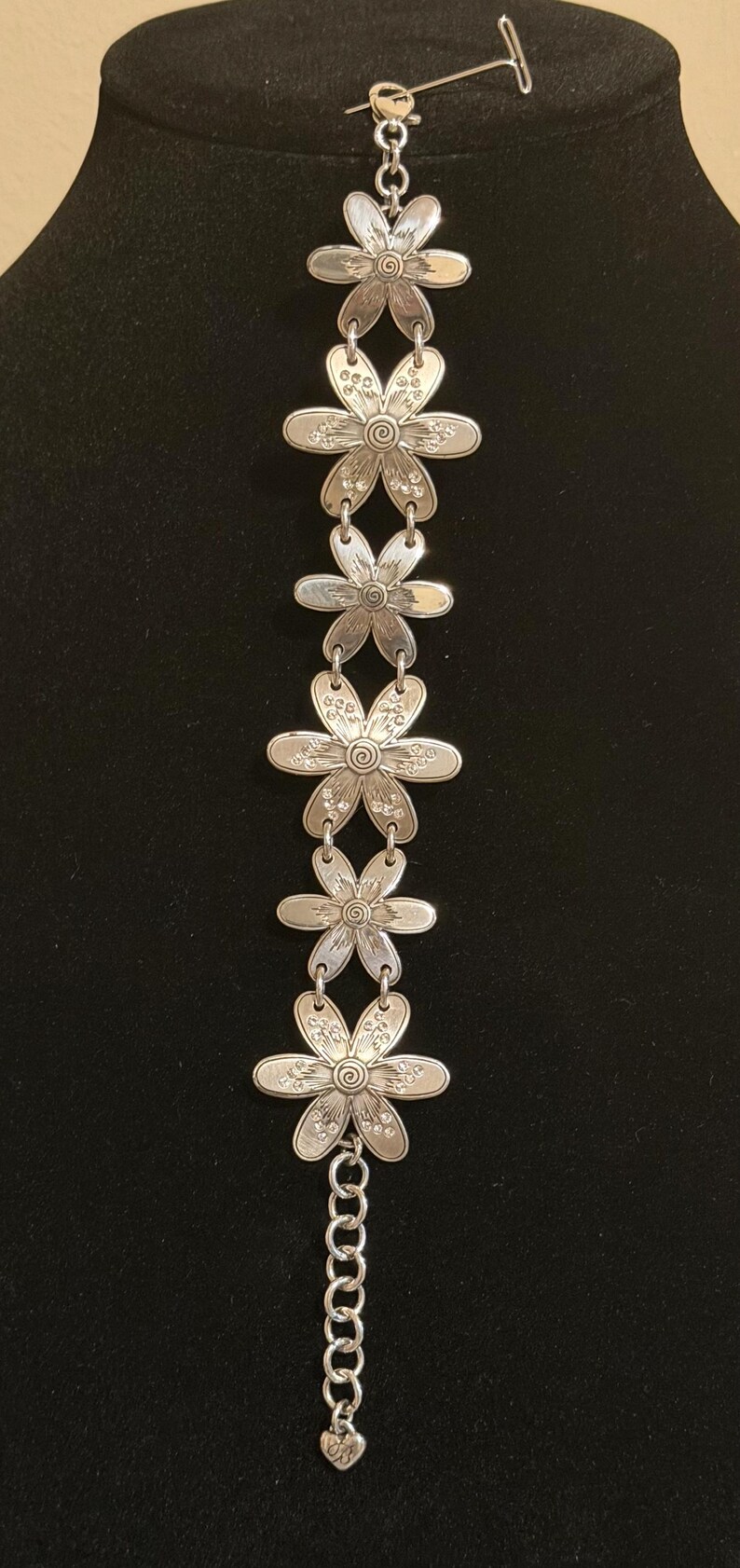 Może przedstawiać: Srebrna bransoletka z ogniwami w kształcie stokrotek. Każdy kwiat ma szczeg&oacute;łowe wzory płatk&oacute;w i środka. Bransoletka jest prezentowana na czarnym tle, podkreślając jej misterne detale.