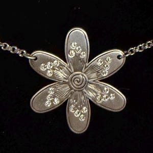 Może przedstawiać: Srebrny naszyjnik z wisiorkiem w kształcie stokrotki. Wisiorek ma sześć płatk&oacute;w z osadzonymi przezroczystymi kryształami i spiralnym środkiem. Łańcuszek jest delikatny i srebrny.