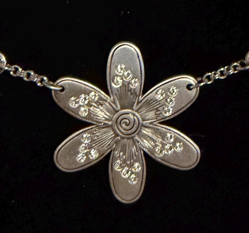 Może przedstawiać: Srebrny naszyjnik z wisiorkiem w kształcie stokrotki. Wisiorek ma sześć płatk&oacute;w z osadzonymi przezroczystymi kryształami i spiralnym środkiem. Łańcuszek jest delikatny i srebrny.