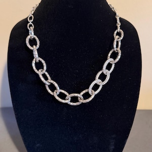 Puede incluir: Un conjunto de collar y pendientes plateados. El collar presenta eslabones ovalados grandes, algunos incrustados con peque&ntilde;os cristales transparentes. Las joyas se exhiben en un maniqu&iacute; negro, mostrando el brillo y el dise&ntilde;o.