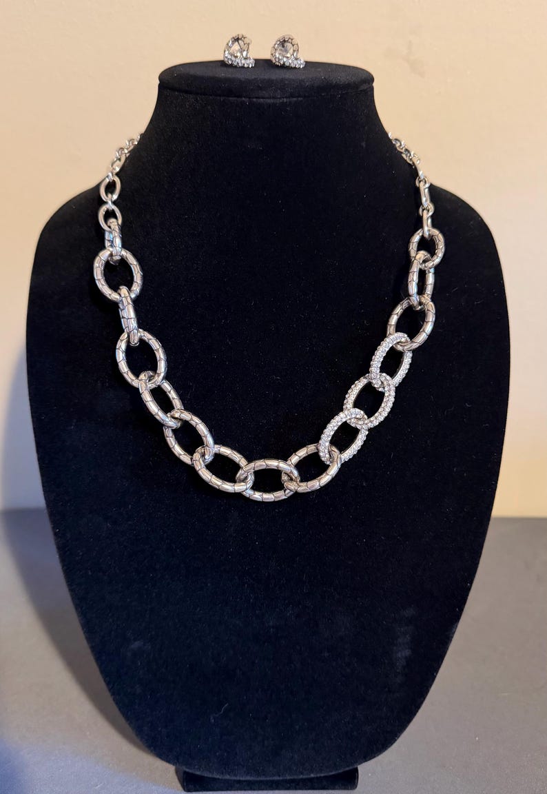 Puede incluir: Un conjunto de collar y pendientes plateados. El collar presenta eslabones ovalados grandes, algunos incrustados con peque&ntilde;os cristales transparentes. Las joyas se exhiben en un maniqu&iacute; negro, mostrando el brillo y el dise&ntilde;o.