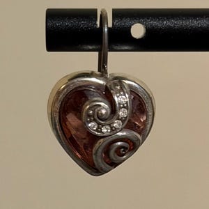 May include: A heart-shaped pendant with a silver-colored frame and a pink center. The pendant features a decorative swirl design and is adorned with small, clear gemstones. The pendant is suspended from a silver-colored hook.
