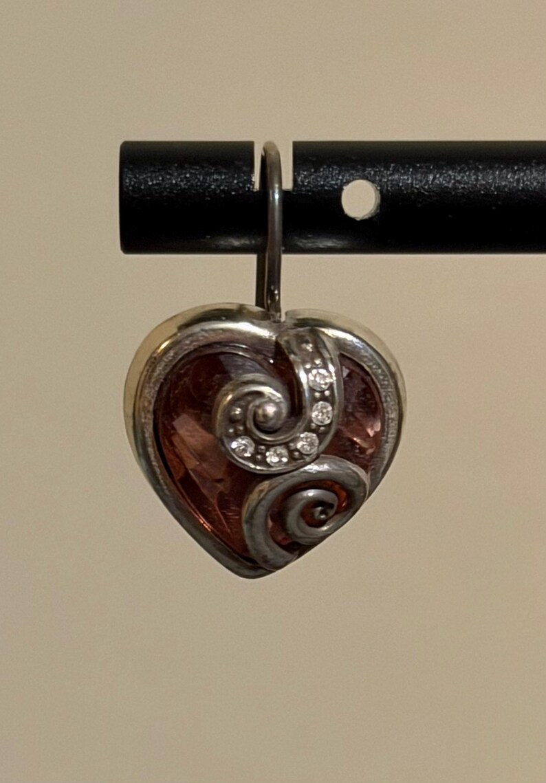 May include: A heart-shaped pendant with a silver-colored frame and a pink center. The pendant features a decorative swirl design and is adorned with small, clear gemstones. The pendant is suspended from a silver-colored hook.