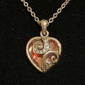 May include: A silver-toned heart pendant necklace with a faceted pink center stone. The pendant features decorative swirls with small clear crystals. The necklace chain is a delicate link style, set against a black background.