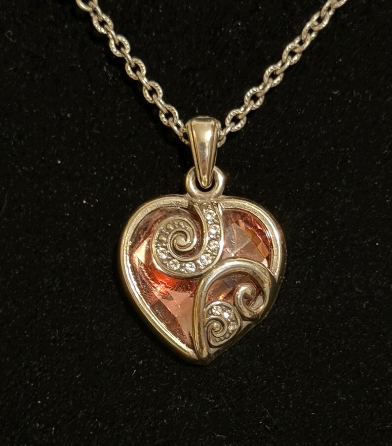 May include: A silver-toned heart pendant necklace with a faceted pink center stone. The pendant features decorative swirls with small clear crystals. The necklace chain is a delicate link style, set against a black background.