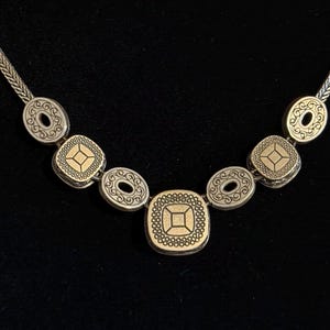 May include: Silver necklace with decorative pendants. The necklace features oval and square-shaped pendants with detailed patterns and gold-coloured accents. The chain has a woven design, and the necklace is displayed against a black background.