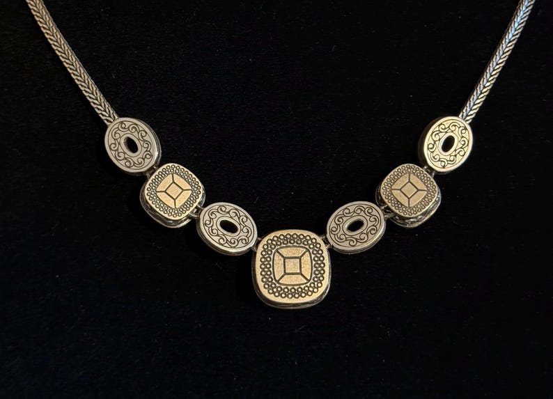 May include: Silver necklace with decorative pendants. The necklace features oval and square-shaped pendants with detailed patterns and gold-coloured accents. The chain has a woven design, and the necklace is displayed against a black background.