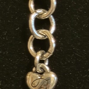 Może przedstawiać: Srebrny łańcuszek z zawieszką w kształcie serca. Zawieszka ma wygrawerowaną kursywą literę "B". Łańcuszek składa się z owalnych ogniw. Tło jest ciemne.