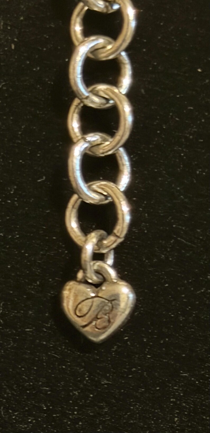Może przedstawiać: Srebrny łańcuszek z zawieszką w kształcie serca. Zawieszka ma wygrawerowaną kursywą literę "B". Łańcuszek składa się z owalnych ogniw. Tło jest ciemne.