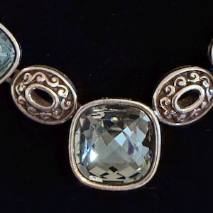 May include: Close-up of a silver necklace with square, faceted, pale green gemstones. The necklace features decorative oval links with an open centre and ornate detailing. The background is black.