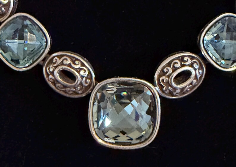 May include: Close-up of a silver necklace with square, faceted, pale green gemstones. The necklace features decorative oval links with an open centre and ornate detailing. The background is black.