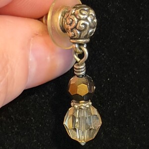 May include: A silver-toned earring with a decorative, swirling design on the post. The earring features two faceted beads, one dark brown and one clear, suspended below the post. The earring is held against a black background.