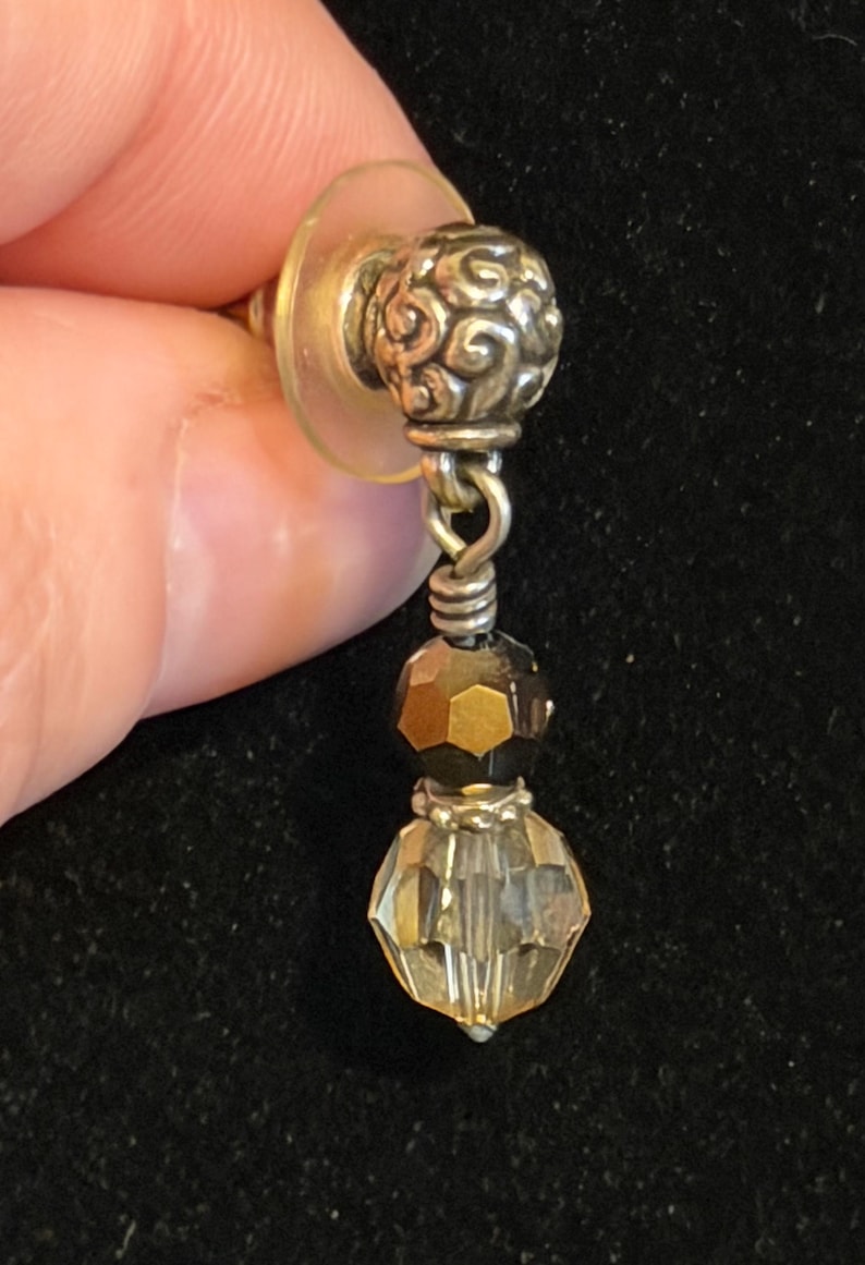 May include: A silver-toned earring with a decorative, swirling design on the post. The earring features two faceted beads, one dark brown and one clear, suspended below the post. The earring is held against a black background.